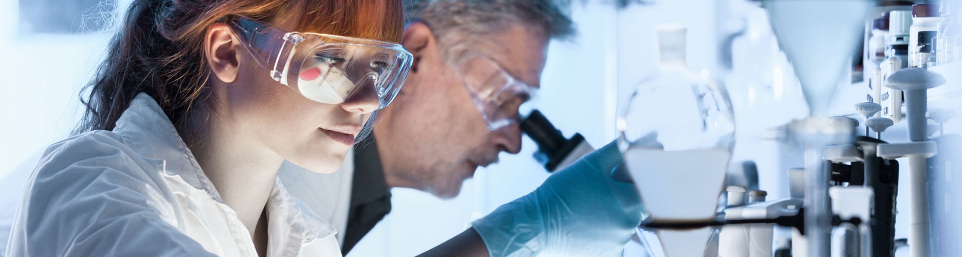 Lächelnde, Brille tragende, Chemie-Laboratin vor einem Hintergrund mit einem Mikroskop und Reagenzgläsern